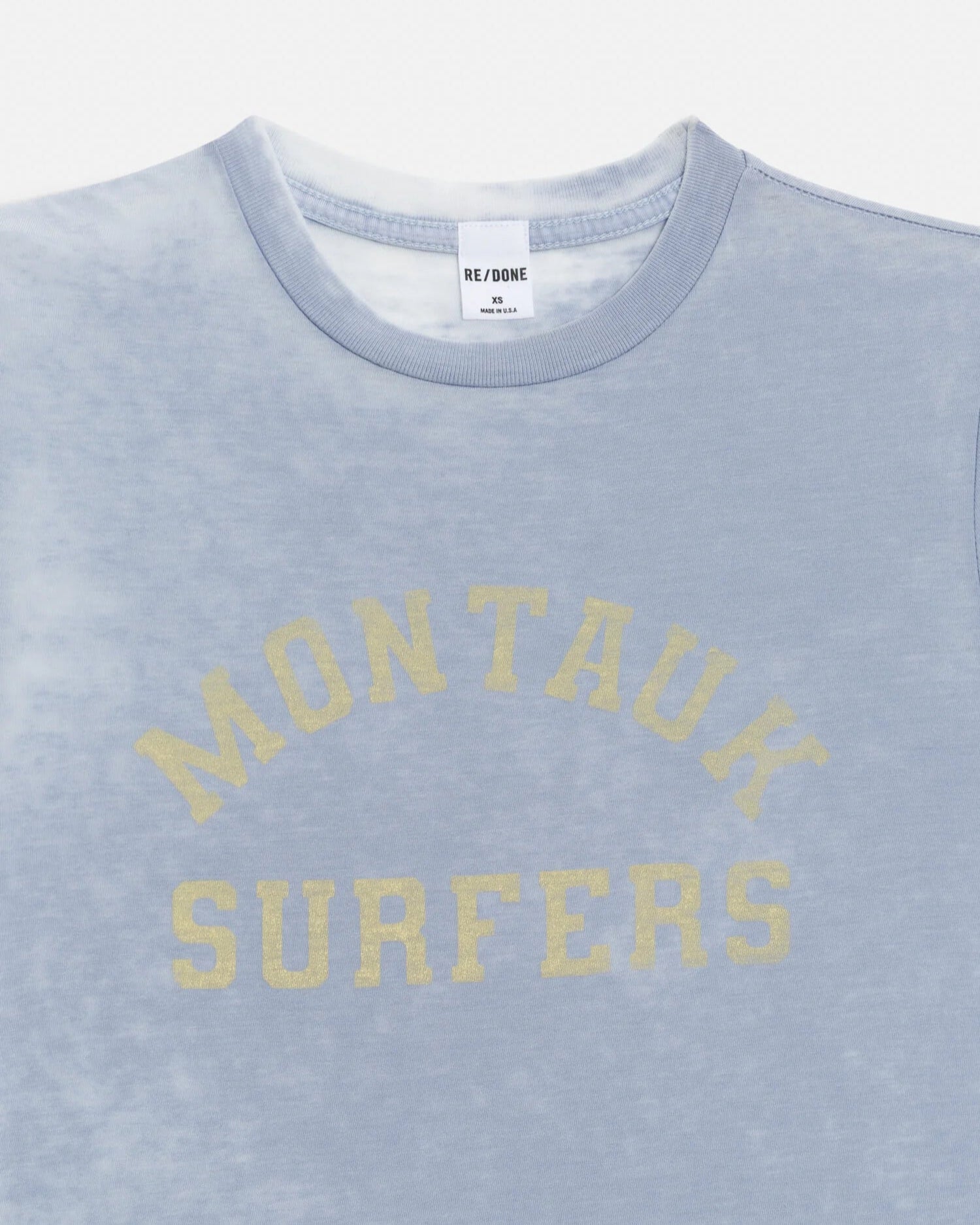 The RE/DONE Montauk Tee is a light blue vintage-style shirt with a faded burnout finish and “MONTAUK SURFERS” in yellow arched text on the chest. The inner tag features the RE/DONE branding.