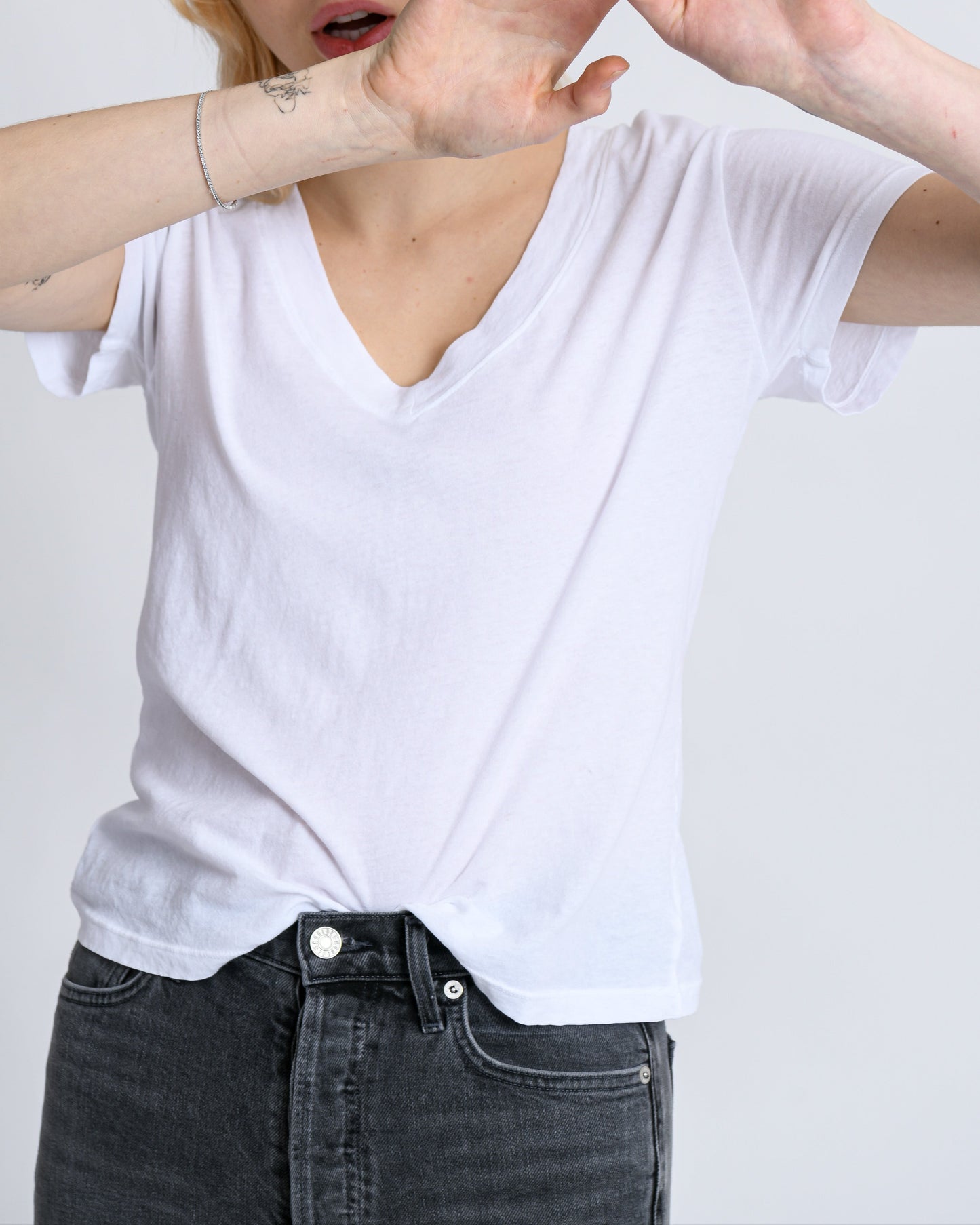 A person with blonde hair in a Hendrix Basic V-Neck T-Shirt White by perfectwhitetee raises their hands in front of their face, appearing in motion against a neutral gray background, showcasing this versatile wardrobe staple.