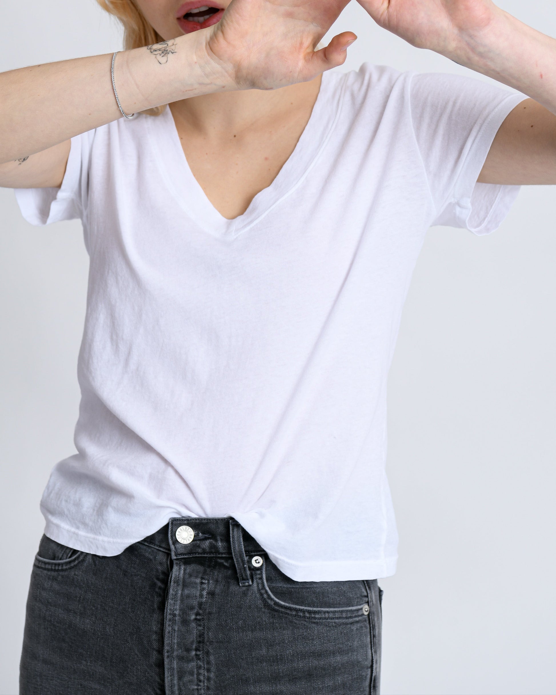 A person with blonde hair in a Hendrix Basic V-Neck T-Shirt White by perfectwhitetee raises their hands in front of their face, appearing in motion against a neutral gray background, showcasing this versatile wardrobe staple.