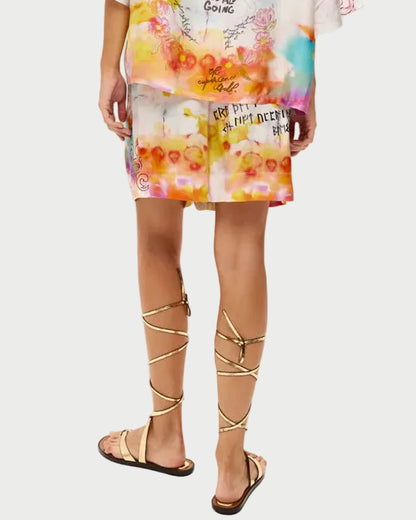 A person wearing ME369 Kori Shorts with colorful, abstract patterns, handwritten text, and artistic designs stands with their back to the camera, paired with gold gladiator-style sandals.