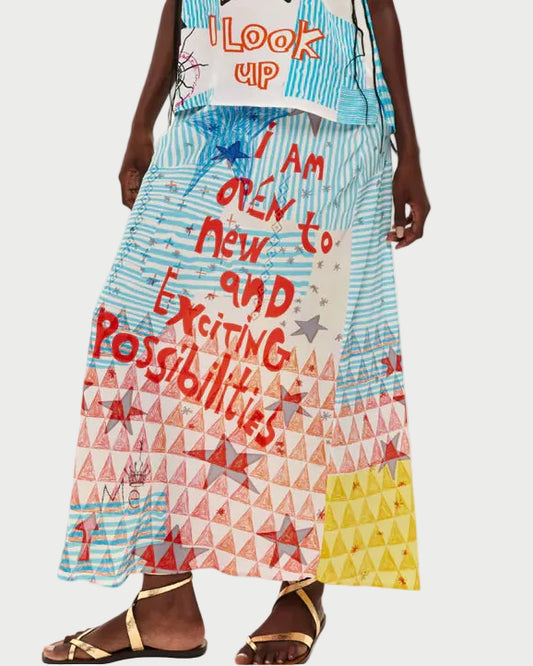 A person wears the ME369 Blakely Slip Skirt by ME369, featuring blue stripes, red and yellow triangles, stars, geometric prints, and the text "I am open to new and exciting possibilities," styled with sandals.
