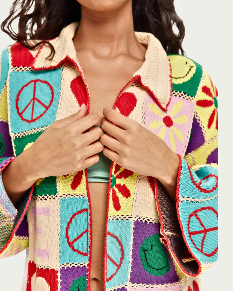 A woman wears the ME369 Miami Crochet Cardigan, featuring colorful granny-square patchwork with peace signs, flowers, hearts, and smiley faces. She holds it partially closed over a mint green top. Her face is mostly out of frame.