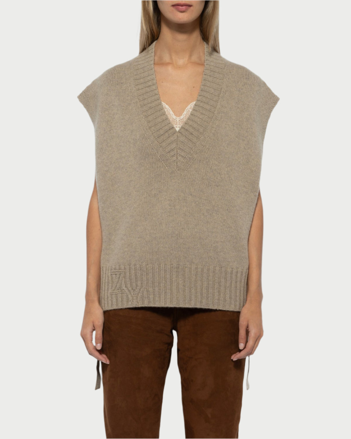 A woman models the Zadig & Voltaire Sweater Vest—a beige, sleeveless knit with a deep V-neck—layered over a lace-trimmed top and paired with brown pants, standing against a white background and looking at the camera neutrally.