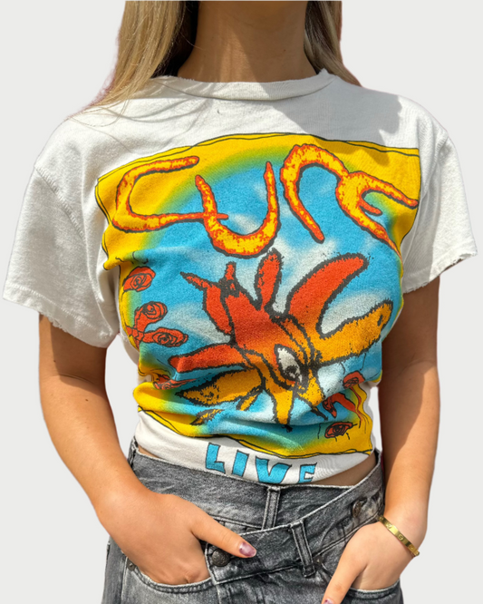 A person wearing a white The Cure Classic Tee by Madeworn with a colorful distressed graphic design stands in front of a red door. The person also wears gray high-waisted jeans and has their hands in their pockets. Their nails are painted in a light purple color.