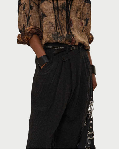 Someone wearing a crinkled brown floral shirt tucked into R13 Triple Pleat Crossover Trousers with a belt, styled with a chunky black bracelet and a black handbag featuring metal details.