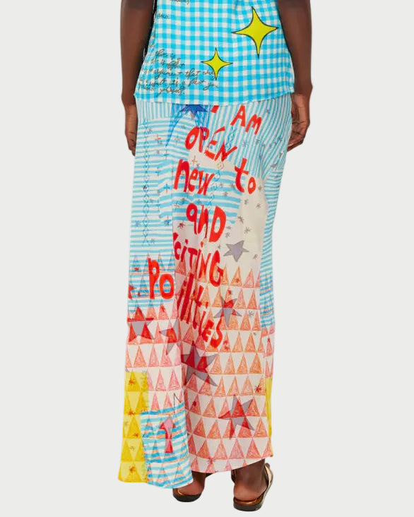 A person wears the ME369 Blakely Slip Skirt by ME369, a colorful maxi slip skirt with blue checkered and red triangle patterns, featuring "I am open to new and exciting possibilities" in bold red letters.
