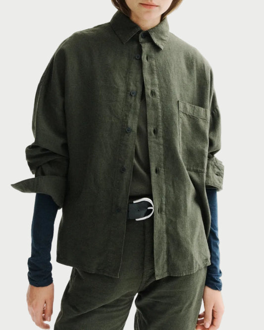 A model wearing an oversized dark olive green linen button-up shirt over a navy blue long-sleeve shirt, styled with matching green trousers and a black belt with a silver buckle, against a plain white studio background.