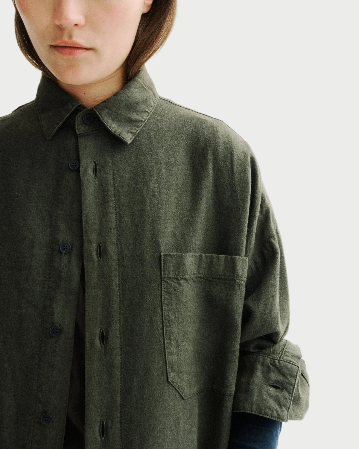 Close-up of a model wearing an oversized dark olive green linen button-up shirt with a chest pocket, set against a plain white background.