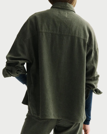 A person shown from the back wearing an oversized dark olive green button-up shirt and matching green pants against a plain light gray studio background.