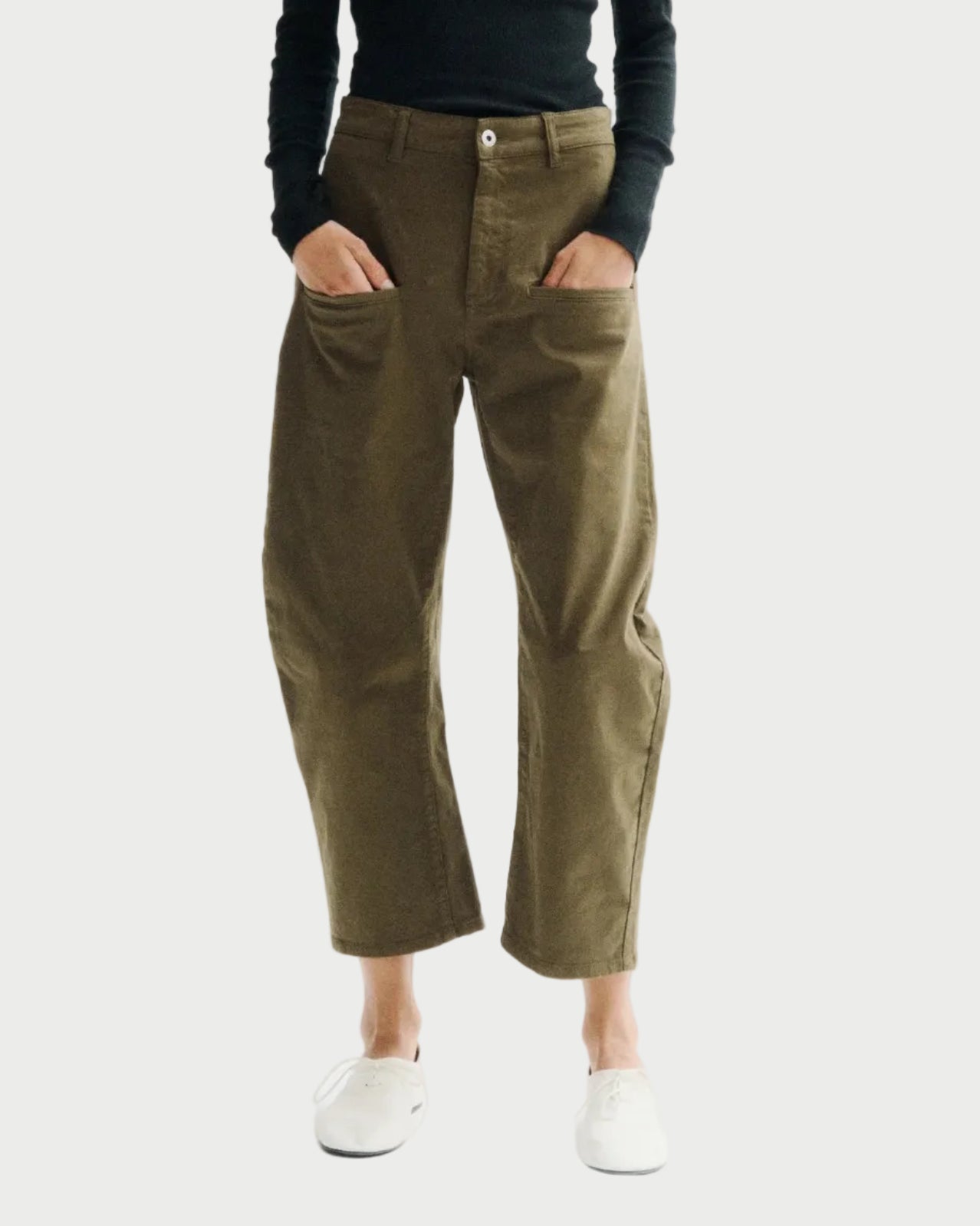 A model stands against a plain white studio background wearing a black long-sleeve turtleneck top, olive green cropped pants, and white sneakers.