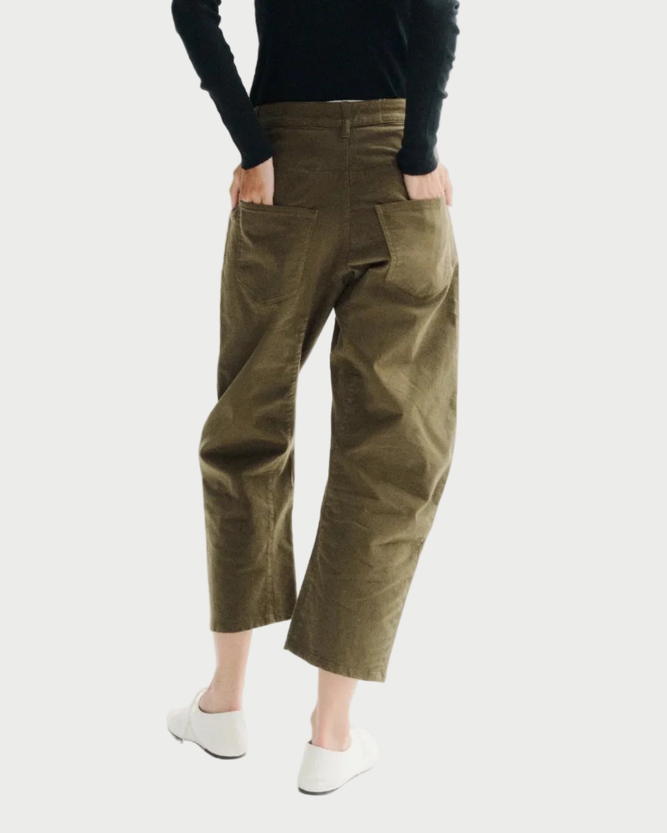 A model viewed from behind wearing olive green cropped trousers, a black long-sleeve turtleneck, and white slip-on shoes against a plain white studio background.
