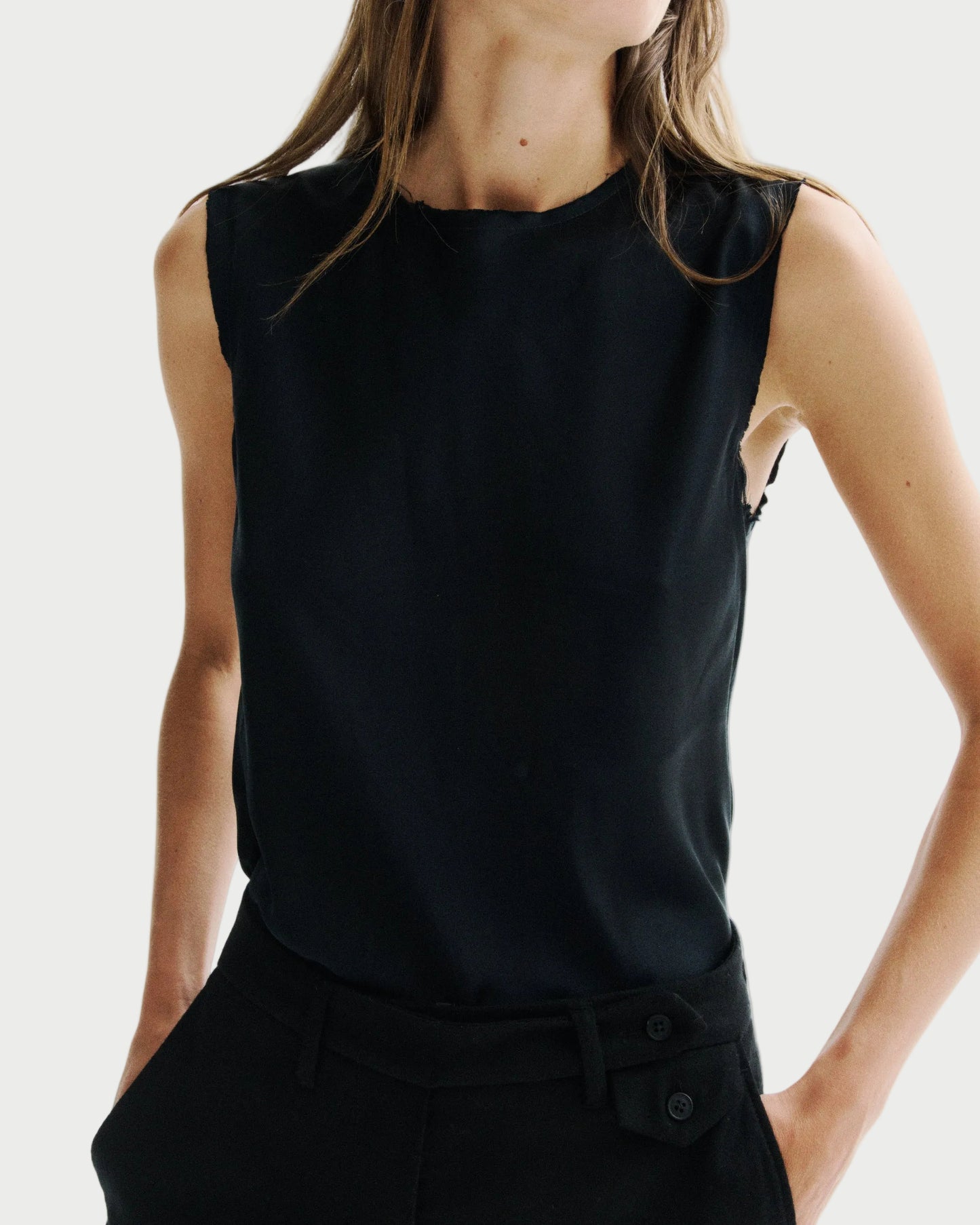 A woman wearing a sleeveless black top and black trousers, cropped from the neck down against a plain light background.