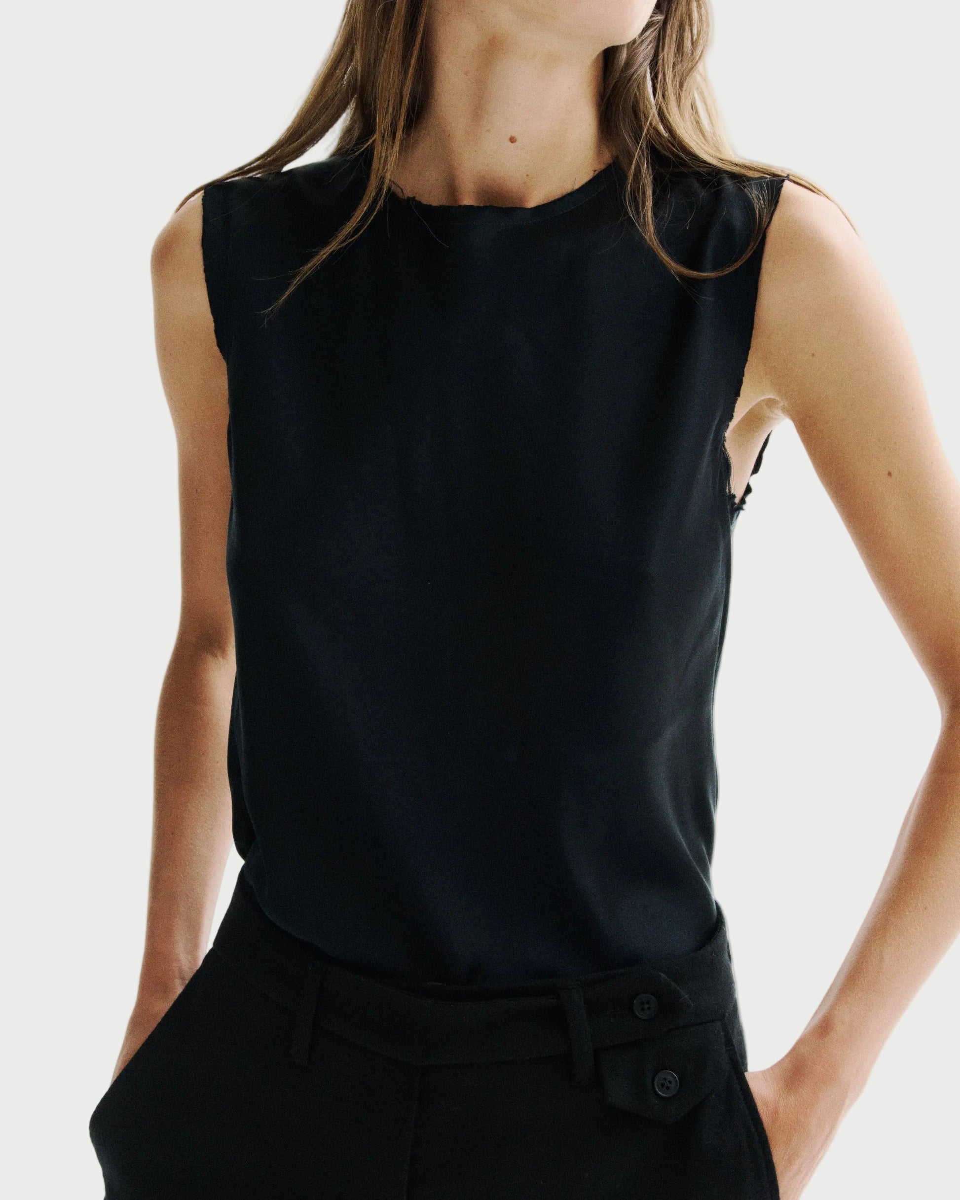 A woman wearing a sleeveless black top and black trousers, cropped from the neck down against a plain light background.