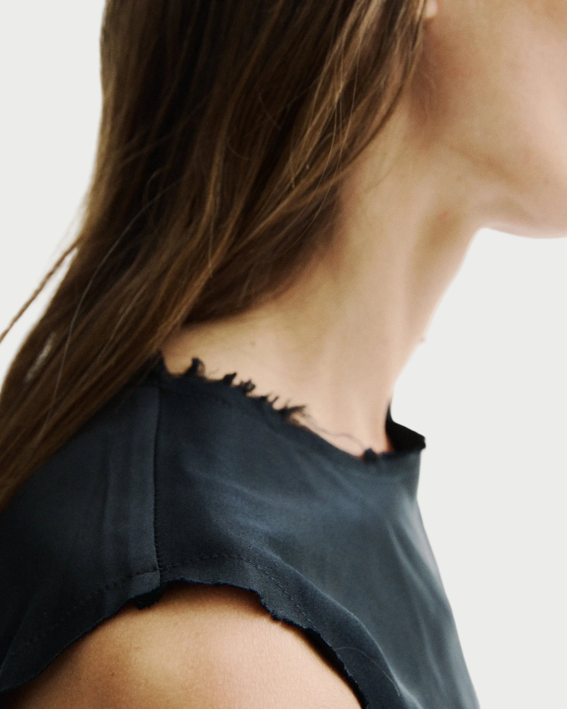Close-up of a woman wearing a dark charcoal sleeveless top with a raw, frayed neckline, shown from the side against a plain white background.
