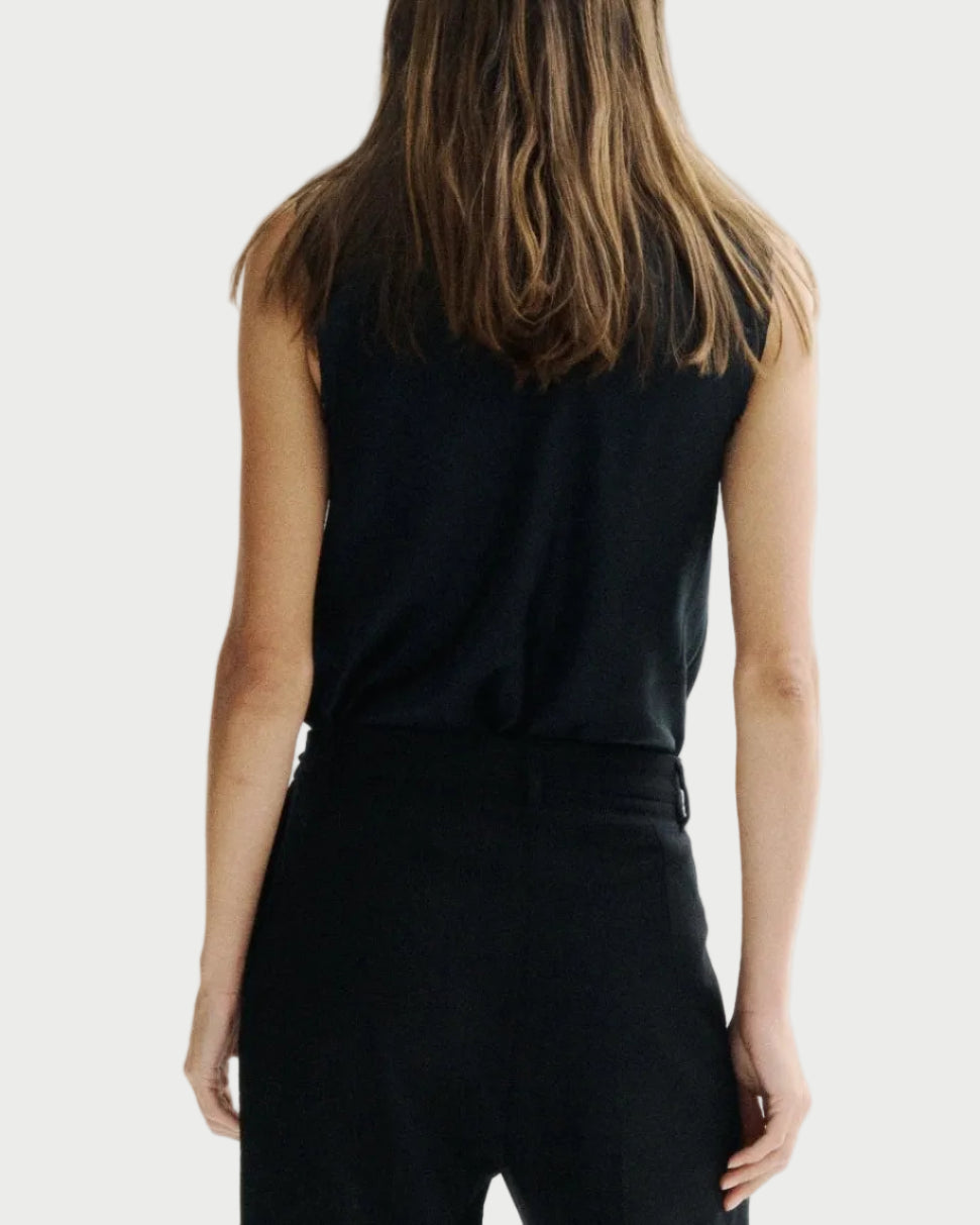 A woman viewed from behind wearing a black sleeveless top and black wide-leg trousers against a plain white studio background.
