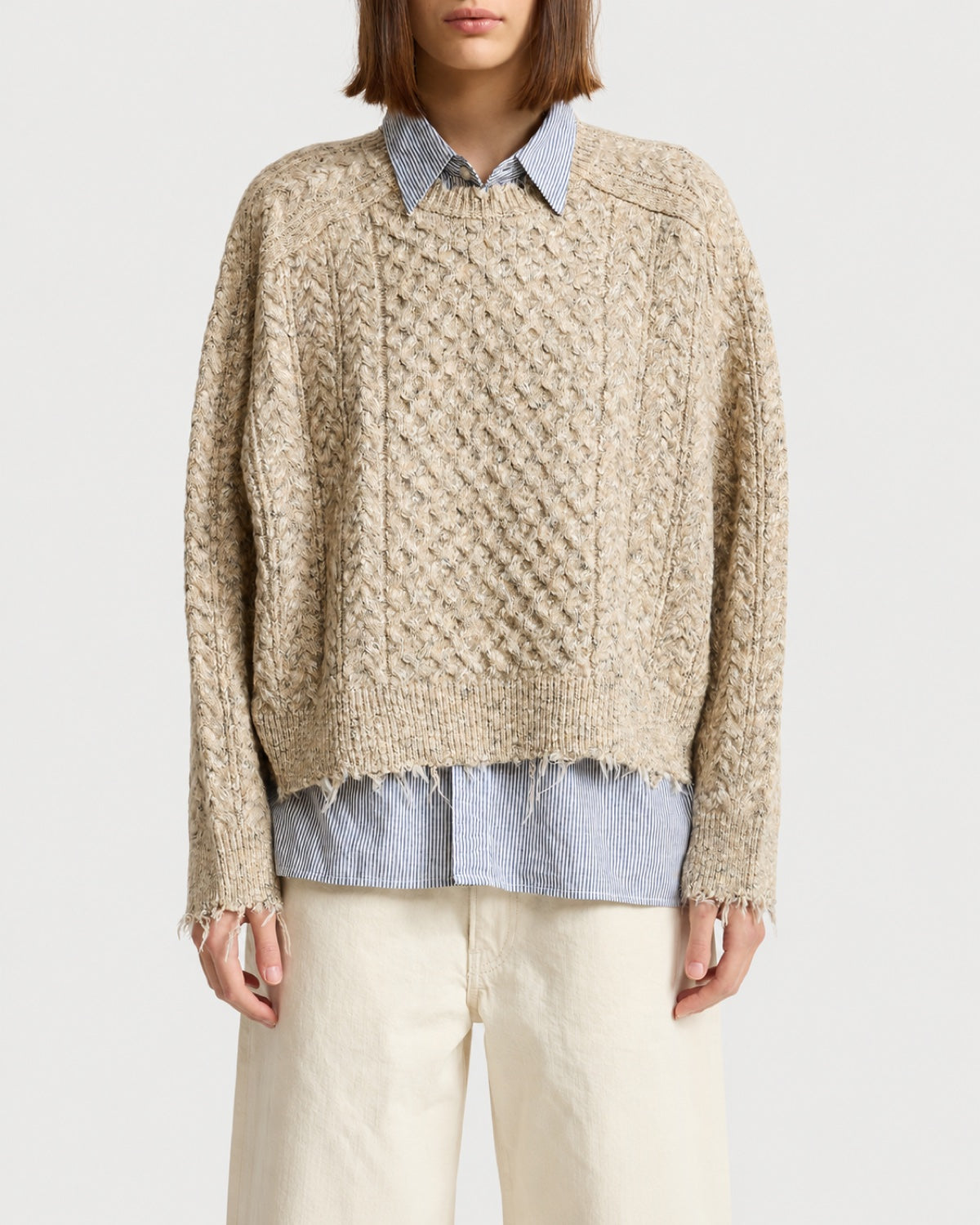A beige Denimist Oversized Cropped Sweater with a boxy fit and a distressed, frayed hem layered over a blue and white striped collared shirt.