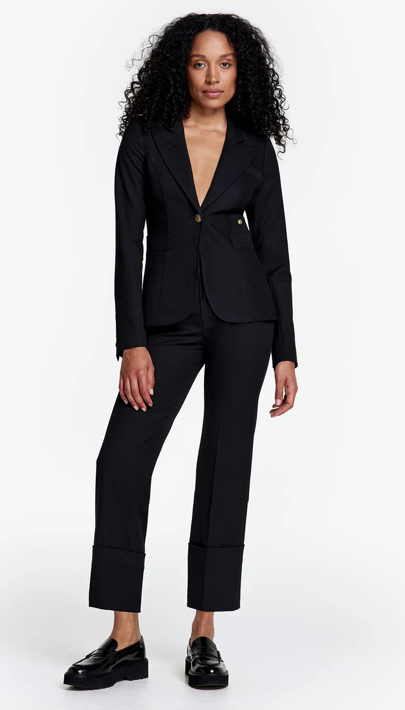 A woman with curly hair wears a Smythe Classic Duchess Blazer in black, styled with cropped, wide-cuffed pants and black loafers, posed against a plain white background.