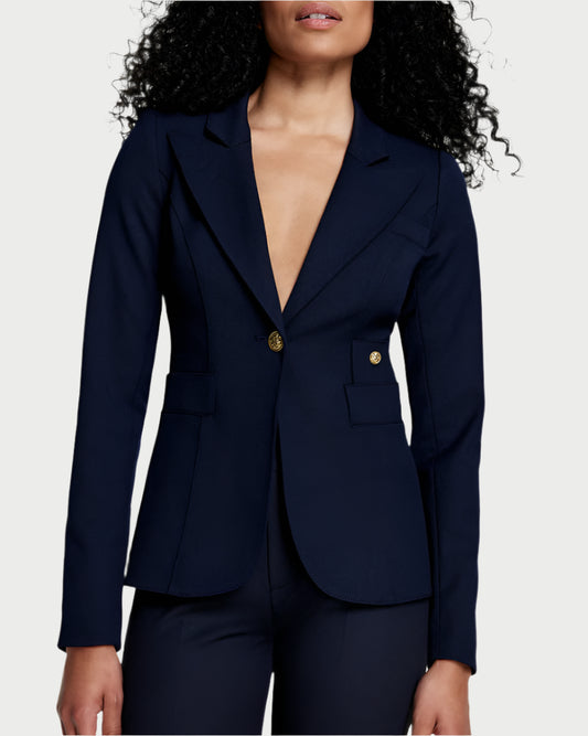 A woman with curly hair wears a Smythe Full Back Duchess Blazer with gold buttons, notched lapels, and flap pockets—no shirt underneath—and matching navy pants against a plain light background.
