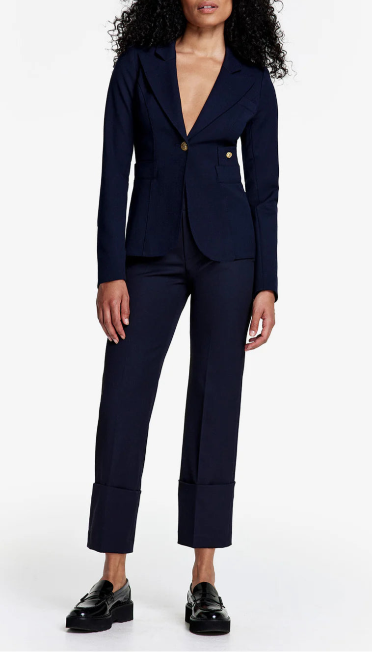 A woman with curly hair models Smythe's Cuffed Flood Pant in Deep Navy with a matching navy blazer and gold buttons. She pairs the outfit with black loafers and stands against a white background.