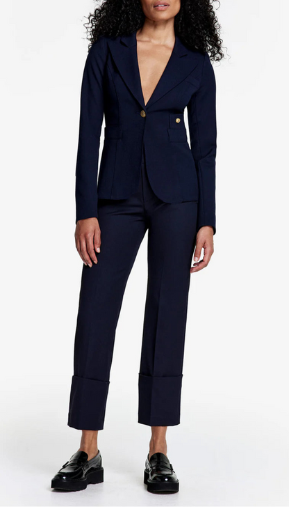 A woman with curly hair models Smythe's Cuffed Flood Pant in Deep Navy with a matching navy blazer and gold buttons. She pairs the outfit with black loafers and stands against a white background.