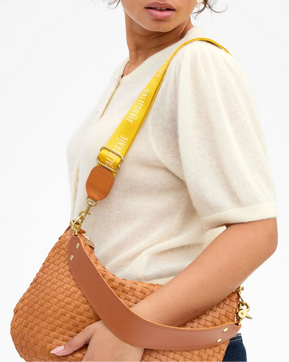 A woman wears a short-sleeved cream sweater and carries a tan woven Clare V. bag with the CLARE V. Californie Adjustable Crossbody Strap, featuring bold yellow with white text to complement its brown leather handle and gold hardware.