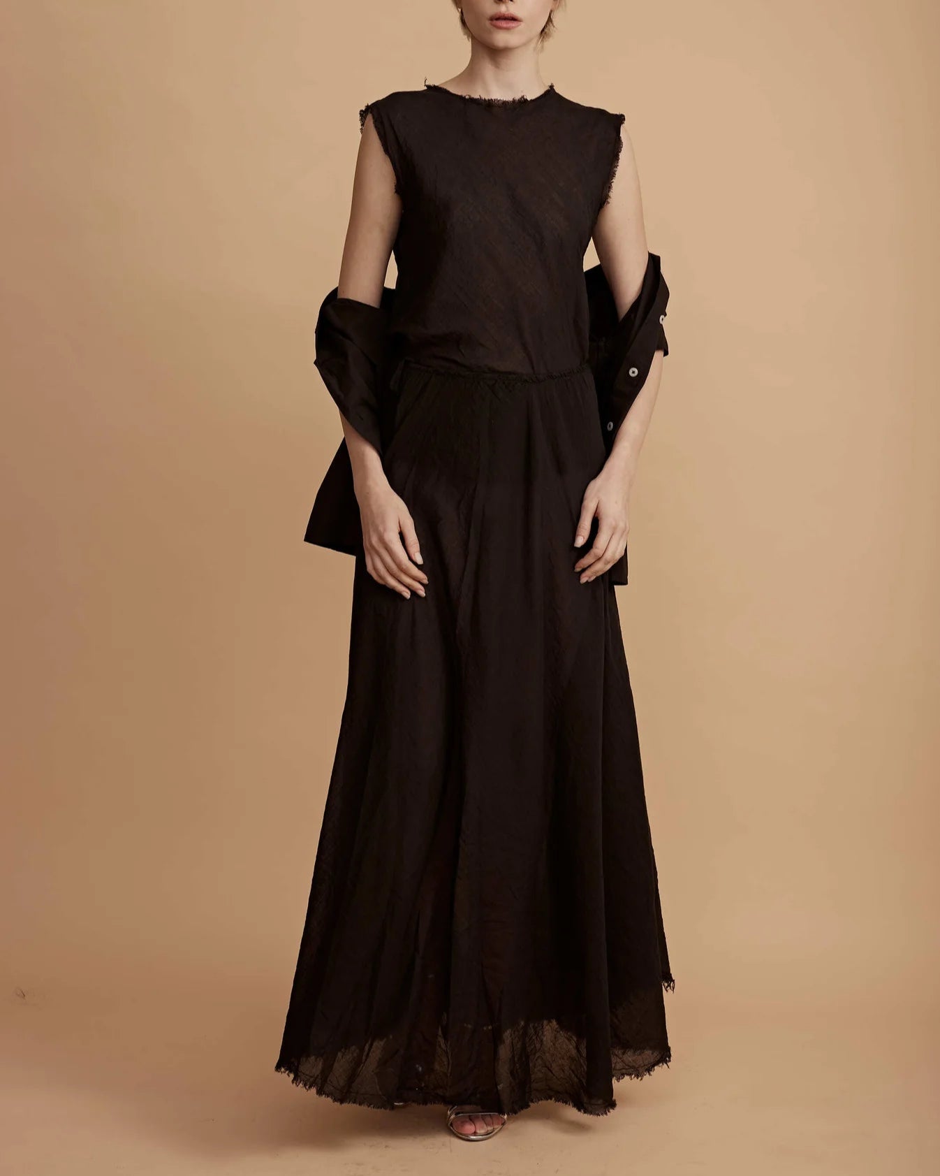 A person stands against a beige background wearing the All III Elysia Maxi Skirt— a flowing cotton style with an elastic waistband—paired with a sleeveless black top and holding a black jacket, their face partially out of frame.