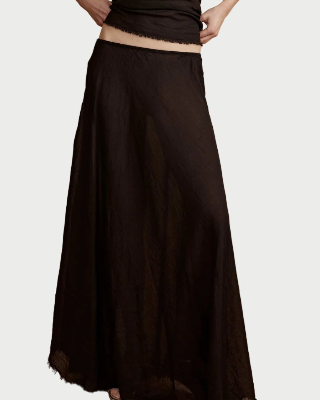 A person wears the All III Elysia Maxi Skirt—a long black semi-transparent skirt with raw hem and elastic waistband—paired with a matching cropped top, standing against a plain light background.