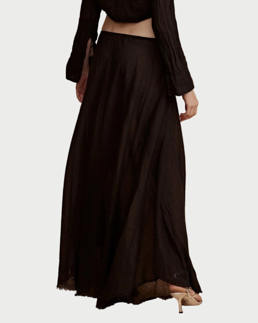 A model is shown from the back wearing the All III Elysia Maxi Skirt in black cotton with an elastic waistband, paired with beige heels and a black long-sleeve top that reveals part of the lower back, set against a plain white background.
