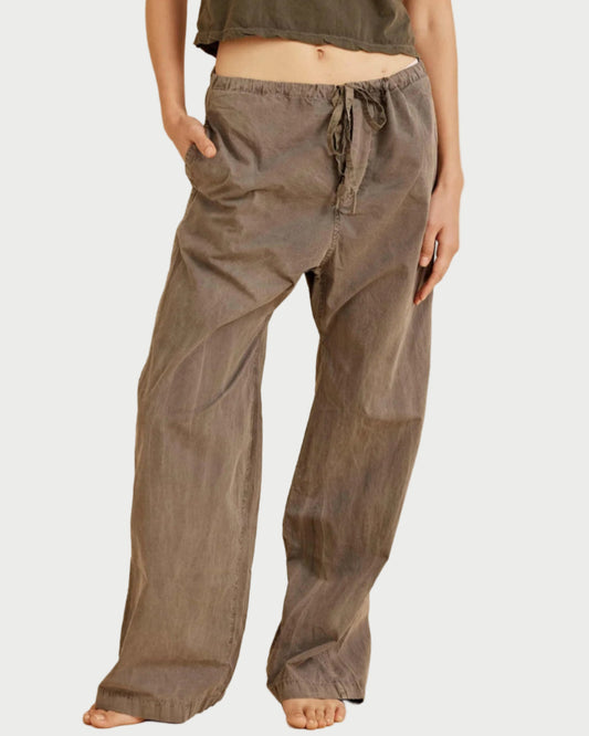 A person stands barefoot, wearing the All III Taylor Pant—a relaxed-fit, brown drawstring utility style—with one hand in their pocket; the upper body is partially visible.
