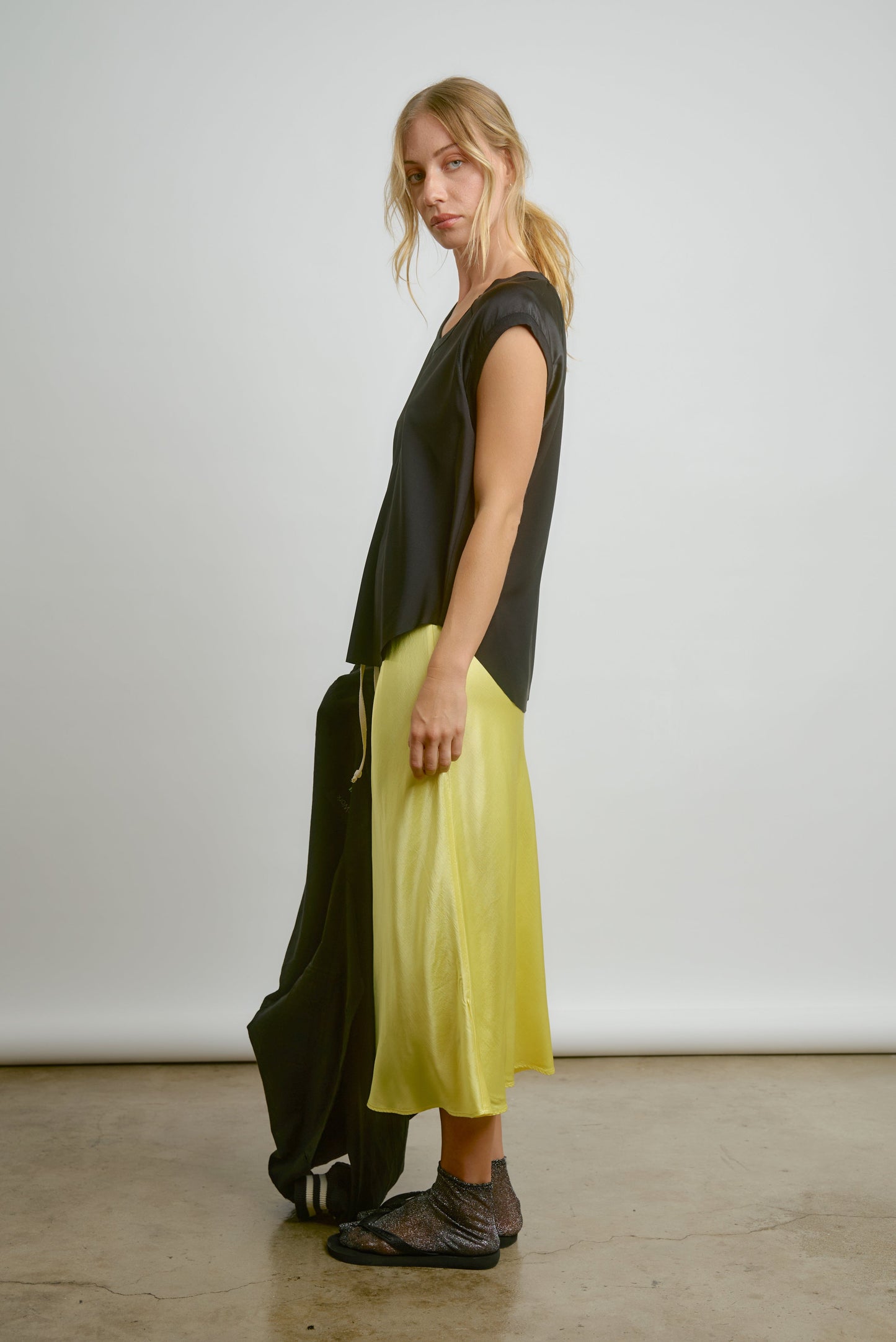 A woman stands sideways against a plain background, wearing the CHAR Cap Sleeve Tee by Aquarius Cocktail, a shiny yellow midi skirt, sheer black socks, black slide sandals, and holding a black jacket. Made in Los Angeles.