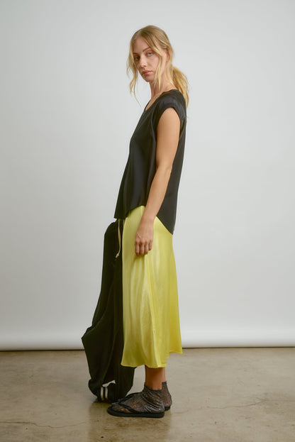 A woman stands sideways against a plain background, wearing the CHAR Cap Sleeve Tee by Aquarius Cocktail, a shiny yellow midi skirt, sheer black socks, black slide sandals, and holding a black jacket. Made in Los Angeles.