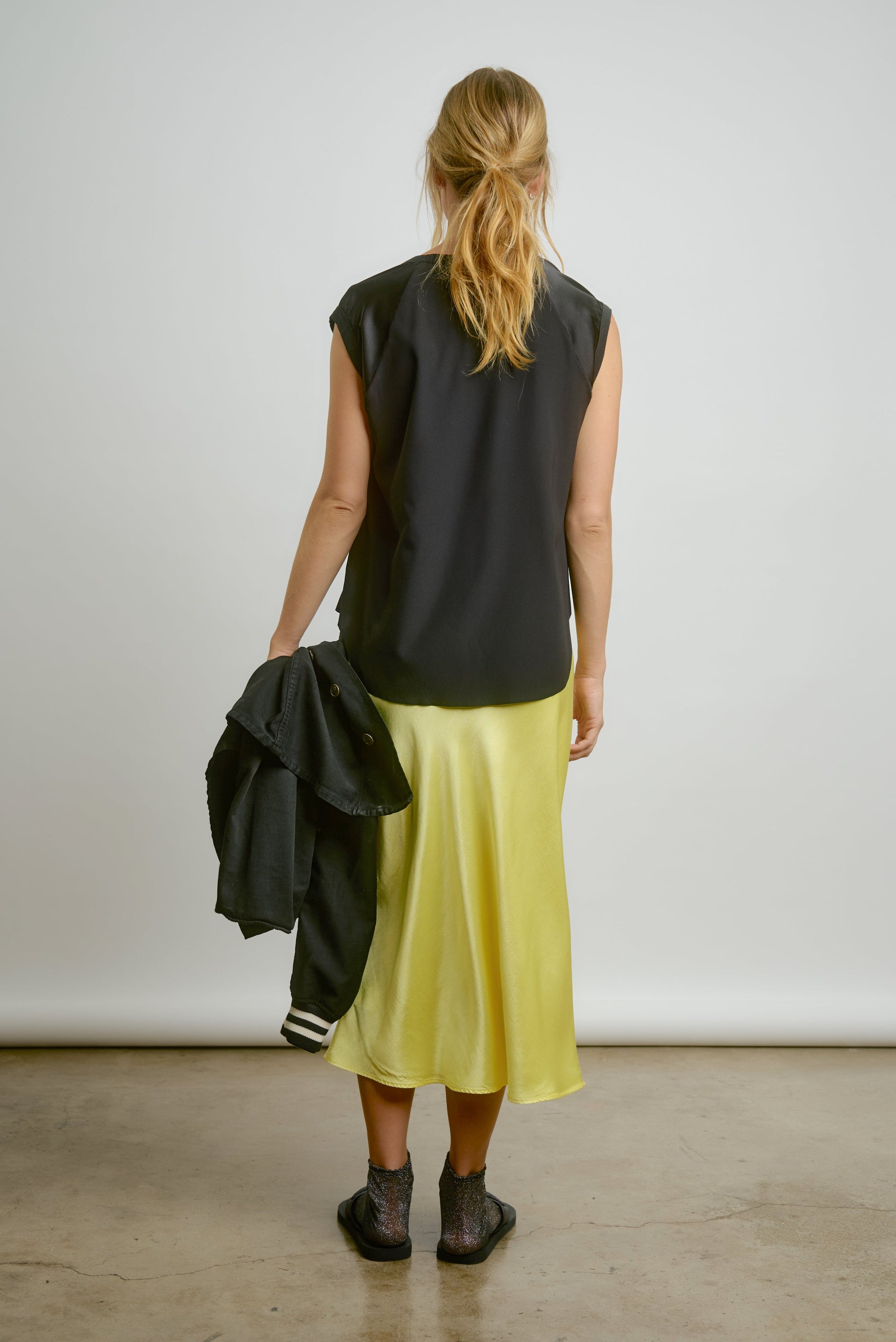 A woman with a ponytail stands on a concrete floor against a plain backdrop, wearing the Aquarius Cocktail CHAR Cap Sleeve Tee in black and a shiny yellow skirt, holding a black jacket in her left hand.