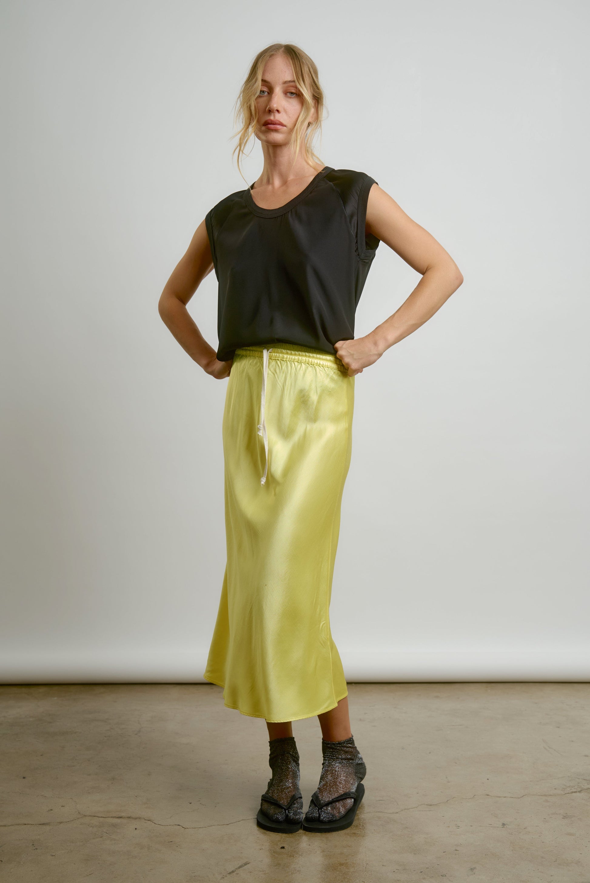 A woman with blonde hair poses confidently against a plain background in the Aquarius Cocktail CHAR Cap Sleeve Tee, paired with a shiny yellow drawstring midi skirt, sheer socks, and black sandals. Made in Los Angeles.