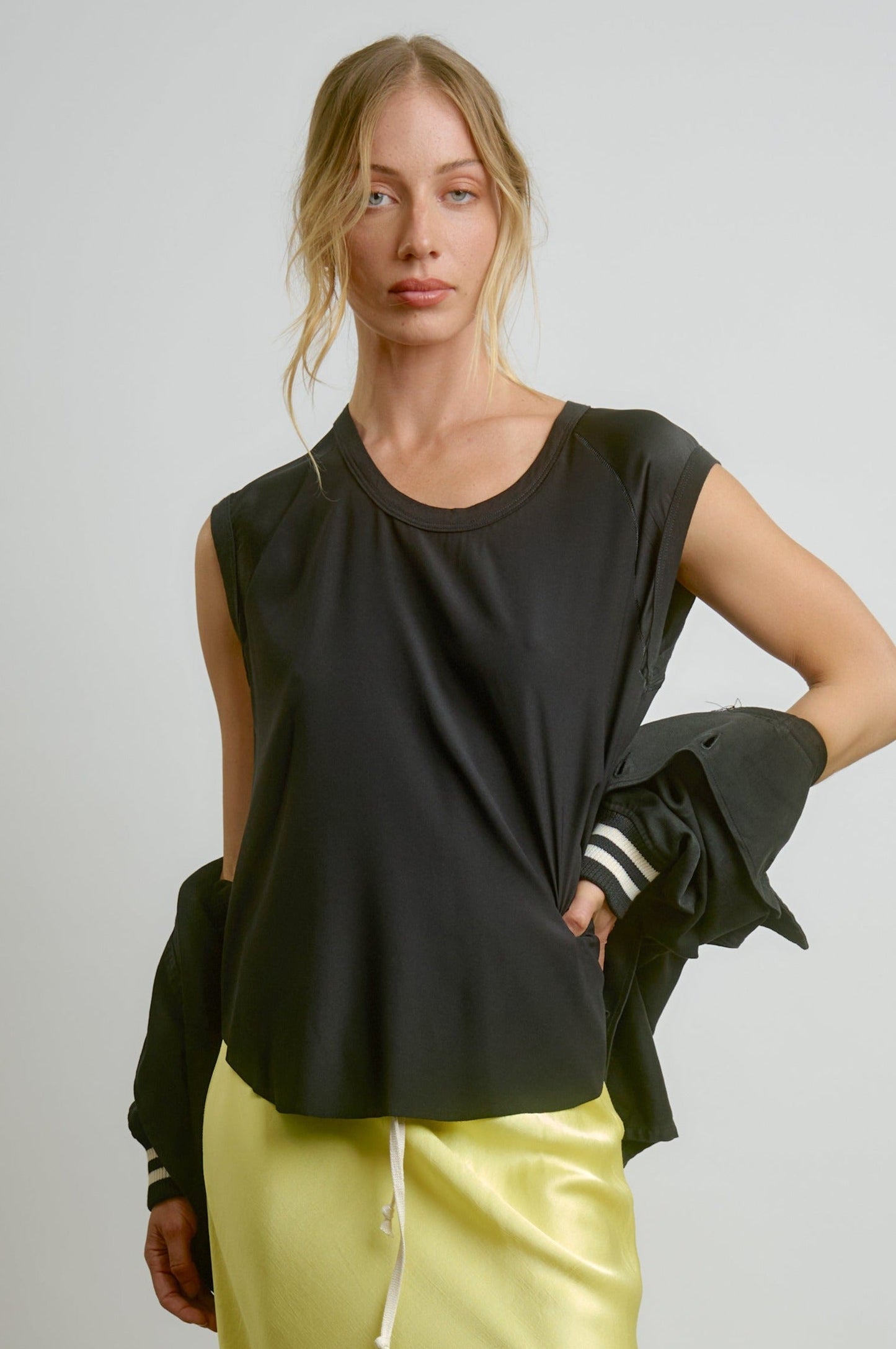 A woman with blonde hair wears the Aquarius Cocktail CHAR Cap Sleeve Tee with a shiny light yellow skirt, a black jacket draped over her arm, standing against a plain light background. The look radiates effortless style, made in Los Angeles.
