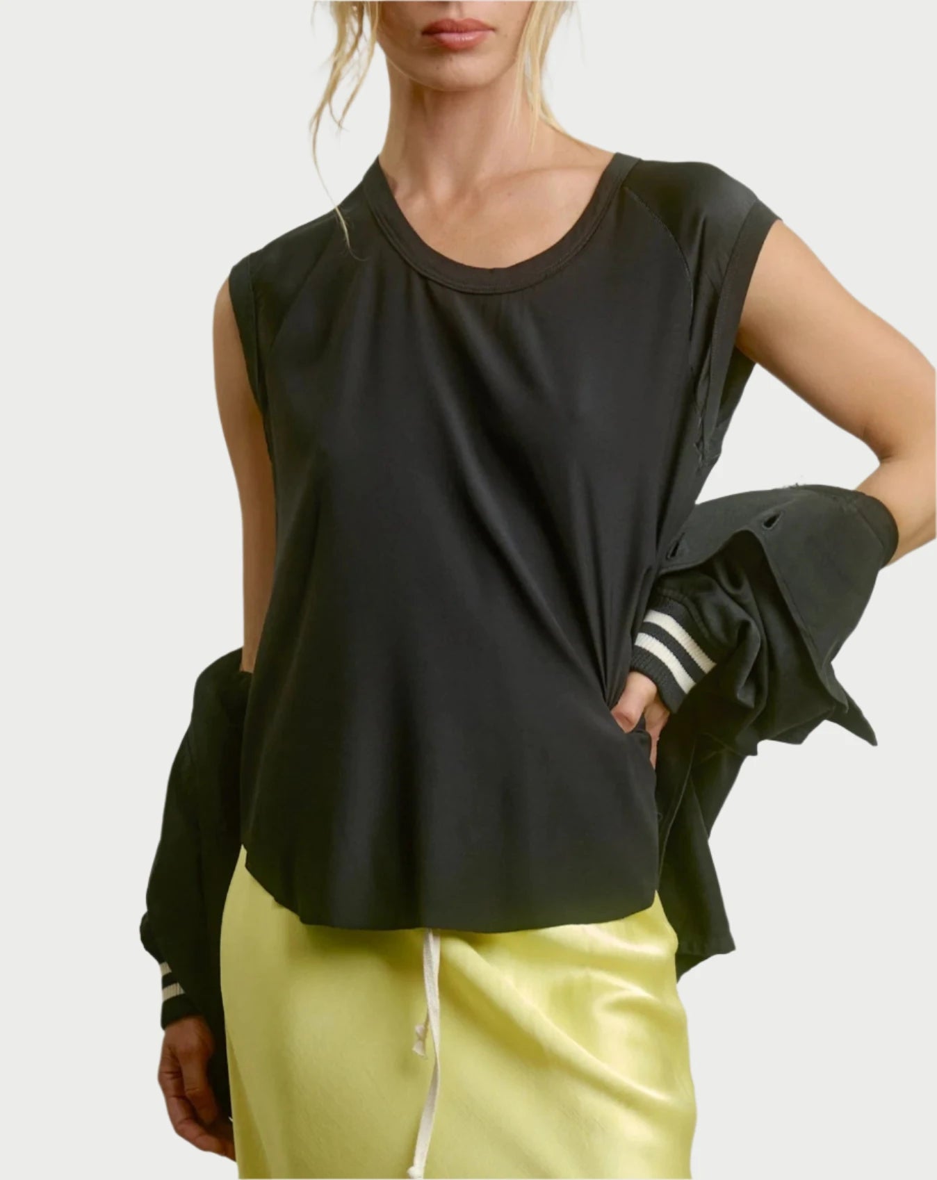 A person with blonde hair poses against a plain background, wearing the Cap Sleeve Baseball Tee in Black by AqC and a pale yellow silk charmeuse skirt. They're holding a dark jacket draped around one arm.