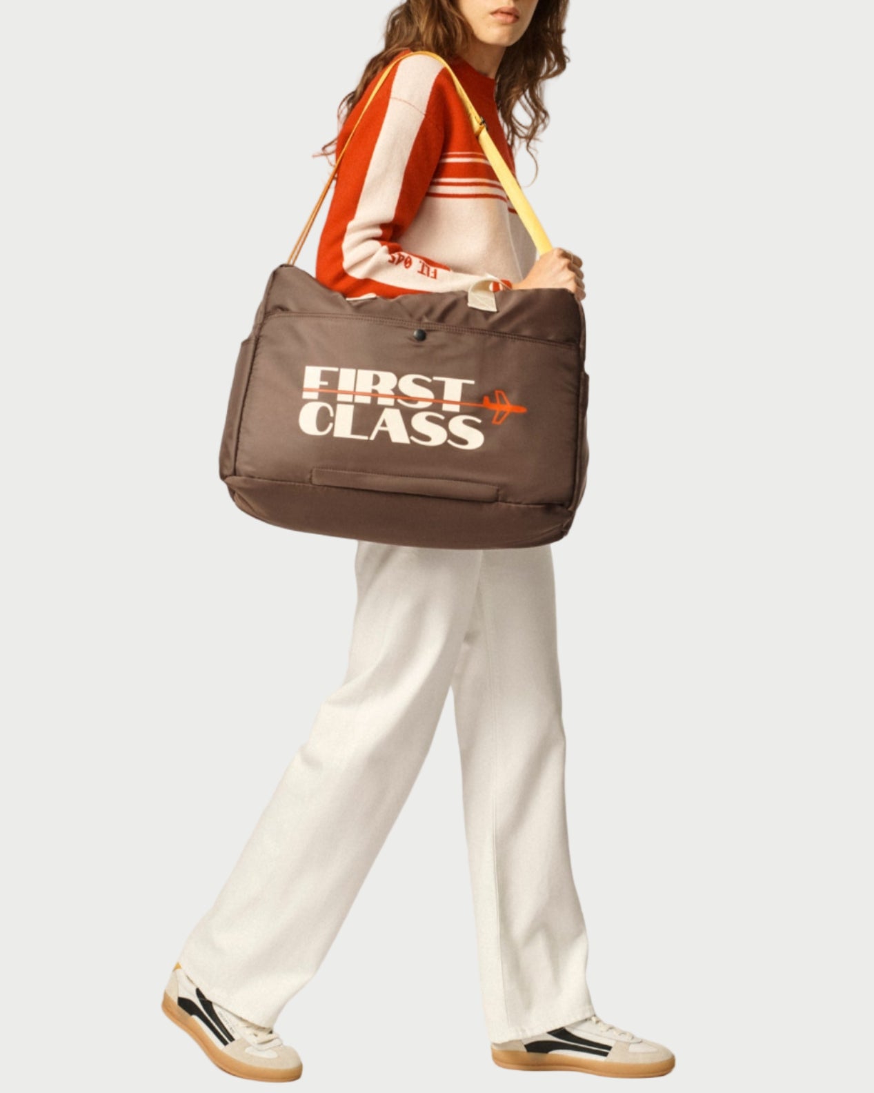 A person wearing a red and white striped sweater, white pants, and sneakers carries the Oncept Olympia Travel Bag by Oncept. The large brown bag features "FIRST CLASS" in white letters. The background is plain light gray.