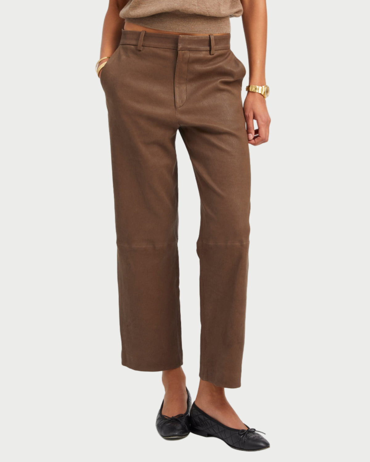 Wearing the SPRWMN Cropped Trousers in Truffle by SPRWMN—crafted from luxurious French lambskin—a person stands with hands in pockets against a plain white background, styled with a brown top, black ballet flats, and a gold watch.