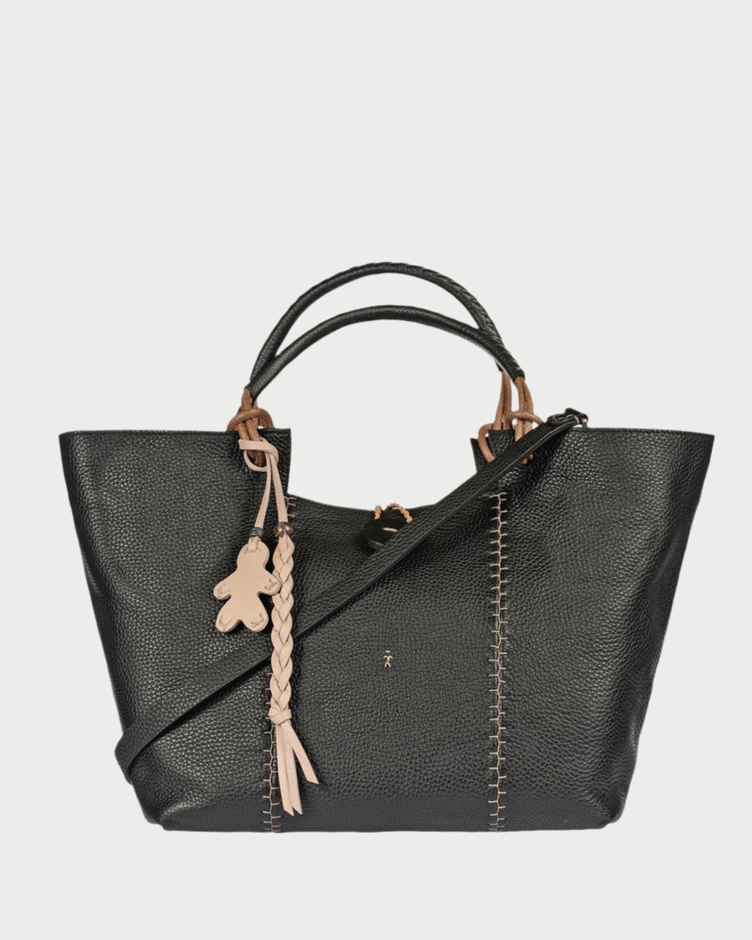 The Medium Muflone Tote by Henry Beguelin is a chic black calfskin leather handbag with tan accents, dual handles, and an adjustable shoulder strap. It includes a teddy bear charm, braided tassel, and prominent front stitching details. Made in Italy.