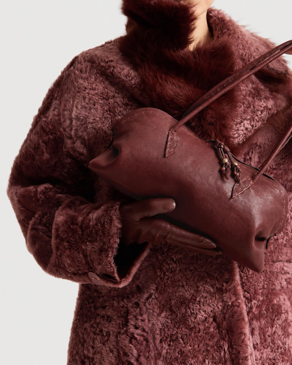 A person wearing a textured burgundy coat and gloves holds a Henry Beguelin Bolla Bassa Bag, a structured, cylindrical luxury handbag in the same deep red shade showcasing fine Italian craftsmanship.