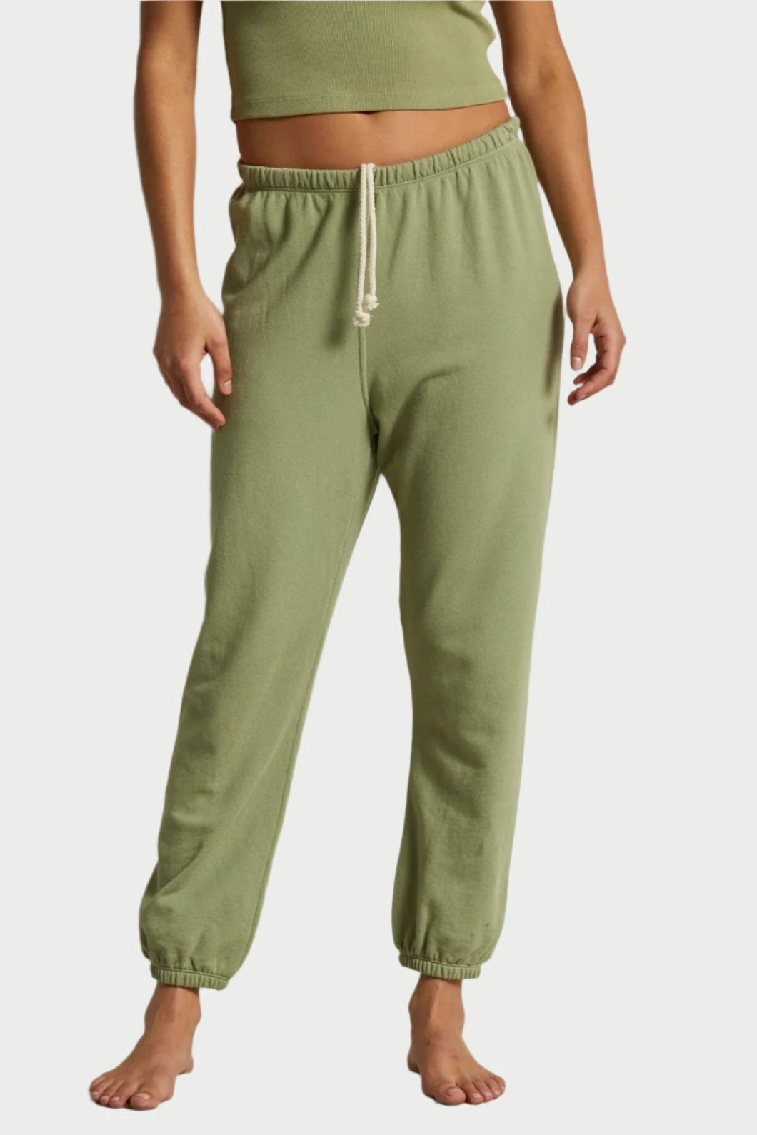 A person stands barefoot on a light-colored draped fabric, wearing a green crop top and matching Johnny Easy Sweatpants from perfectwhitetee, made from buttery soft French terry cotton. Their arms are relaxed at their sides, and the sweatpants have elastic cuffs at the ankles.