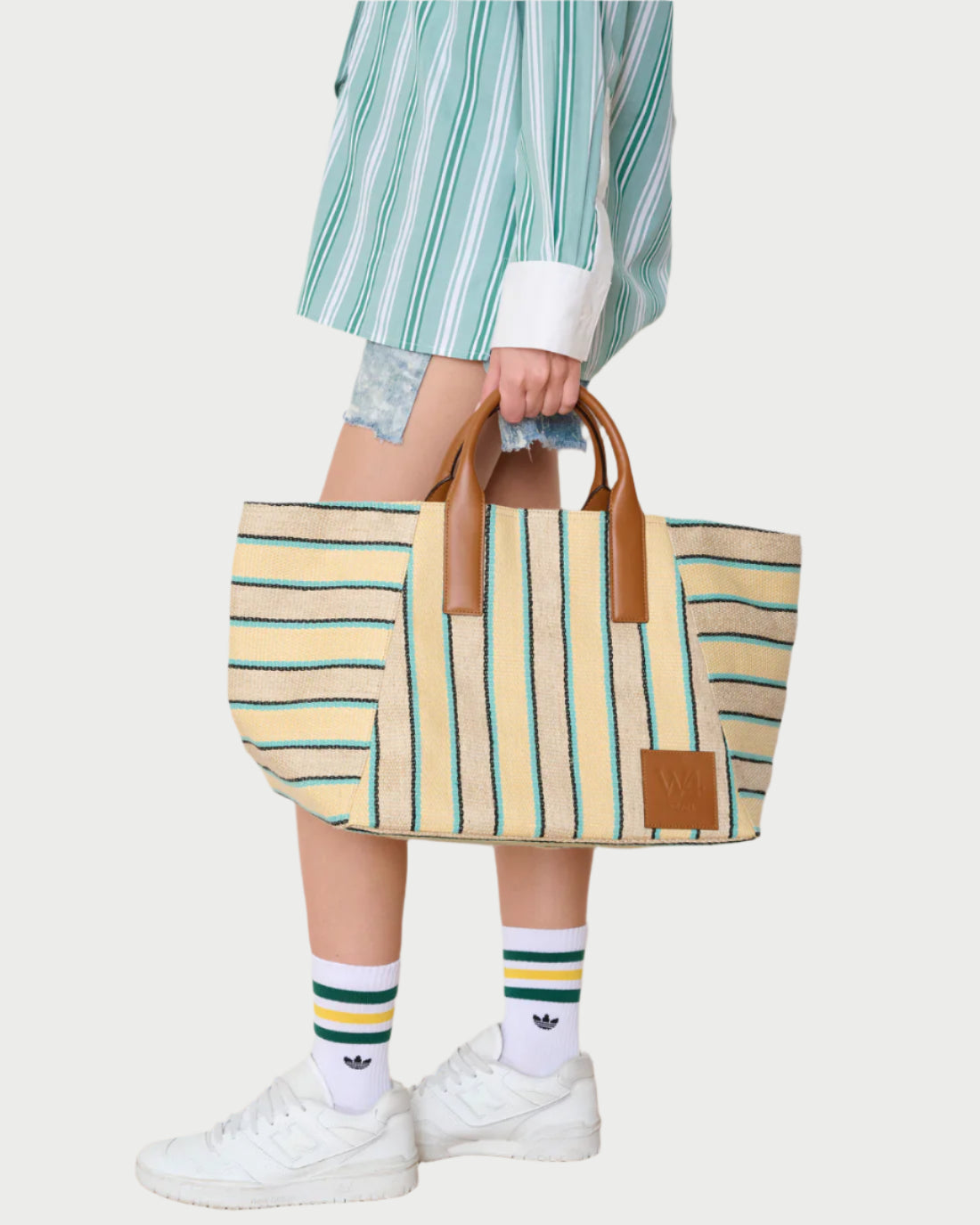 A person in a green striped shirt, denim shorts, and white Adidas sneakers carries the WE-AR4 Riviera Tote—a beige canvas bag with green stripes and brown leather handles.