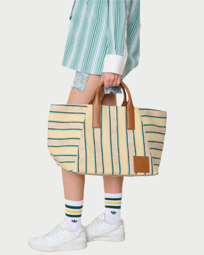 A person in a green striped shirt, denim shorts, and white Adidas sneakers carries the WE-AR4 Riviera Tote—a beige canvas bag with green stripes and brown leather handles.