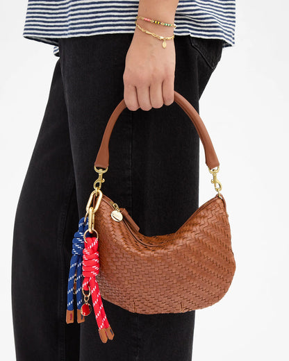 A person wearing a striped shirt and black pants holds a small, brown woven handbag with a curved handle, adorned with red and blue keychains—including the Clare V. Barrel Knot Fob by CLARE V.—as an accessory. The person also wears a gold bracelet with small charms.