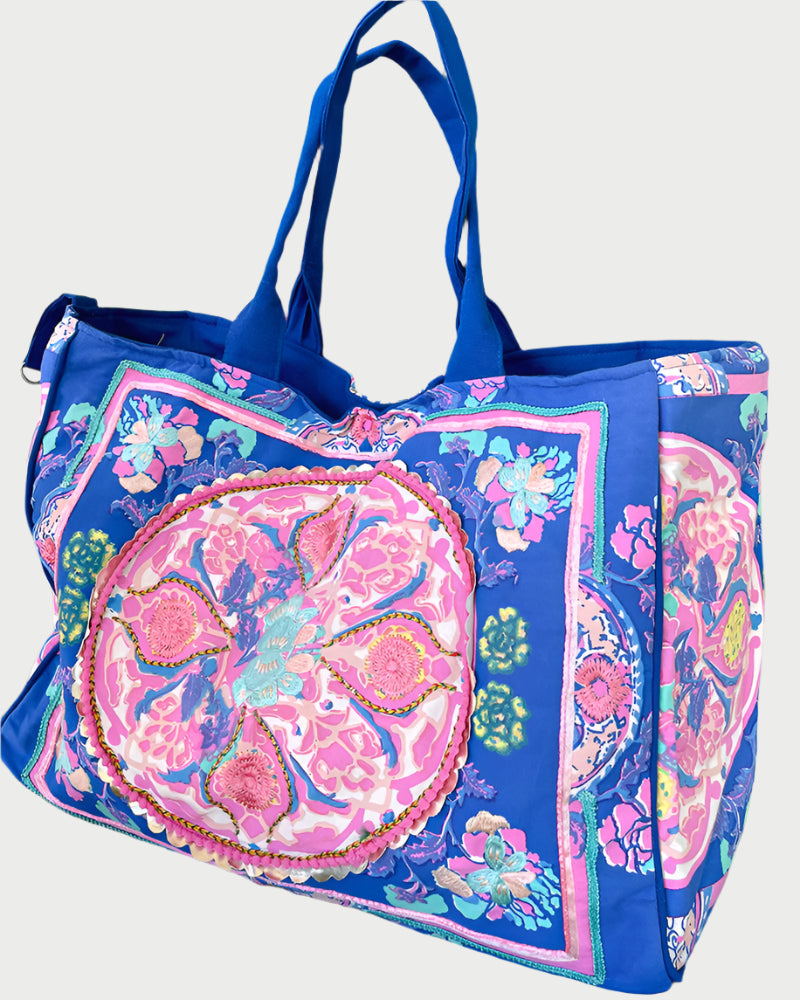 The DEBBIE KATZ Boho Bag in vibrant blue showcases colorful, intricate patterns with pink and green floral motifs, resembling artisanal embroidery. Its lively design is ideal for beach days and comes with sturdy blue handles for durability.