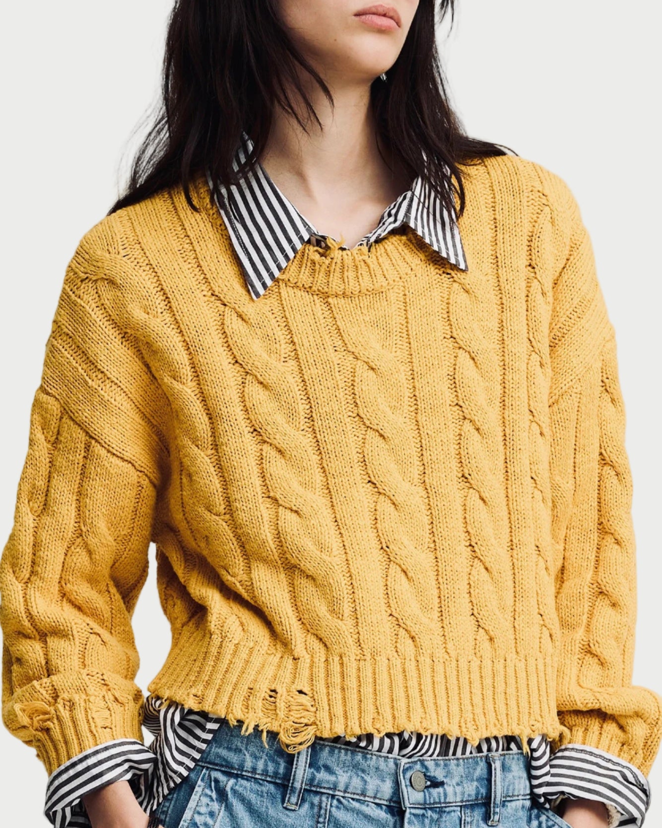 Person wearing a mustard yellow cable knit sweater over a striped shirt with a plain background