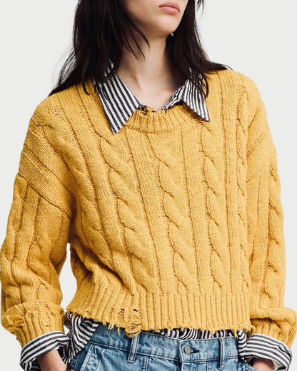 Person wearing a mustard yellow cable knit sweater over a striped shirt with a plain background