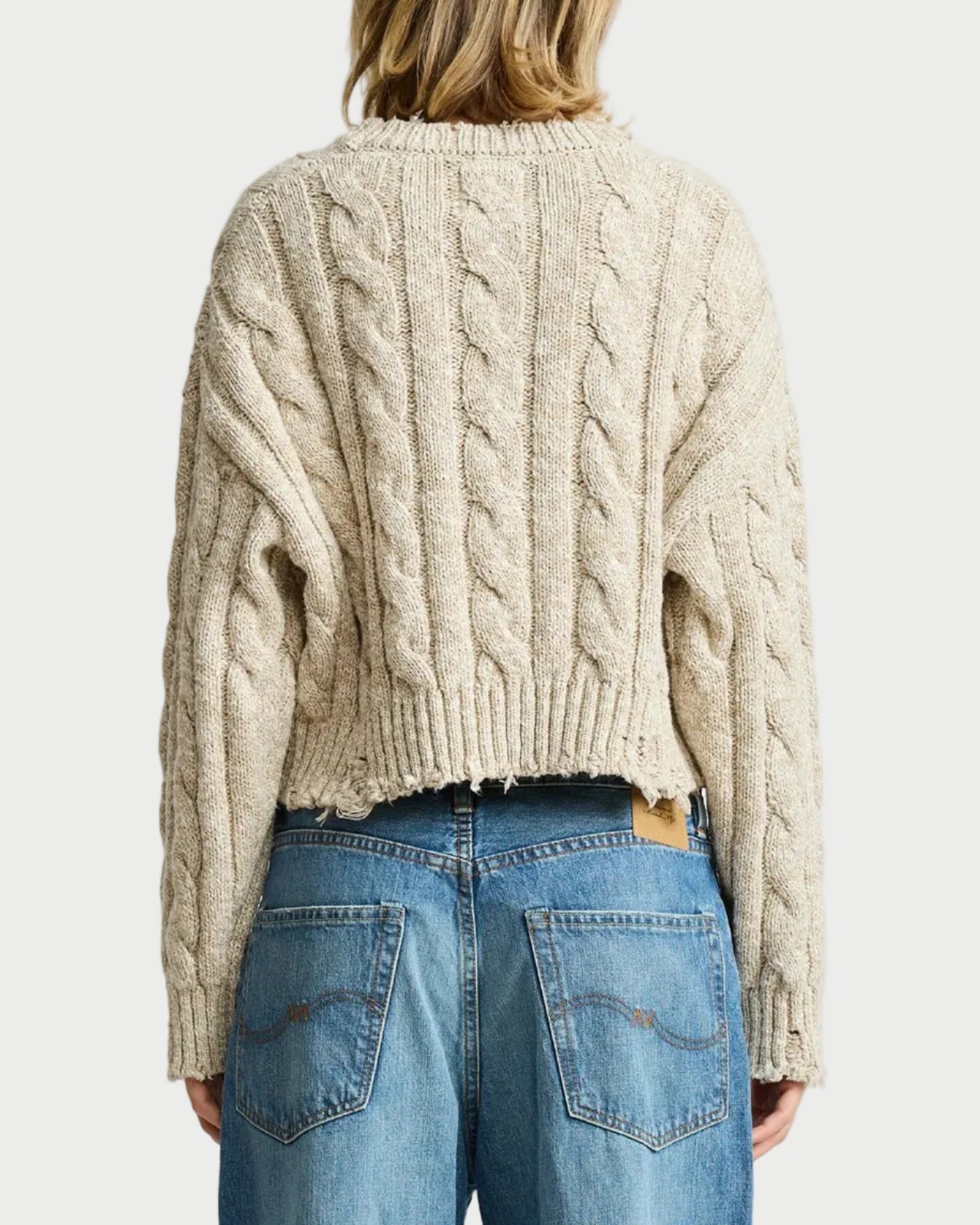 A woman viewed from the back wearing a cream cable-knit cropped sweater and blue jeans against a light gray studio background.