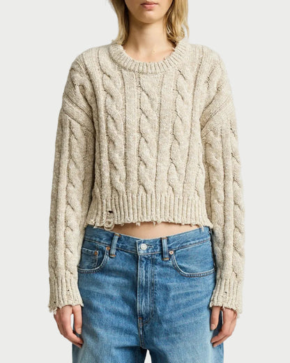 A woman wearing a cropped, cable-knit beige sweater and blue jeans, standing against a light gray studio background.