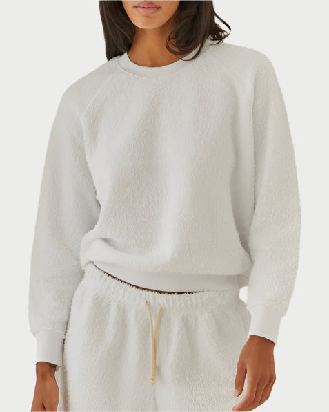 A woman stands against a plain white background wearing the perfectwhitetee Sami Boucle Sweatshirt, a cozy, lightweight top with raglan sleeves. Her matching fleece pants feature a drawstring waist, and her face is partially out of frame.