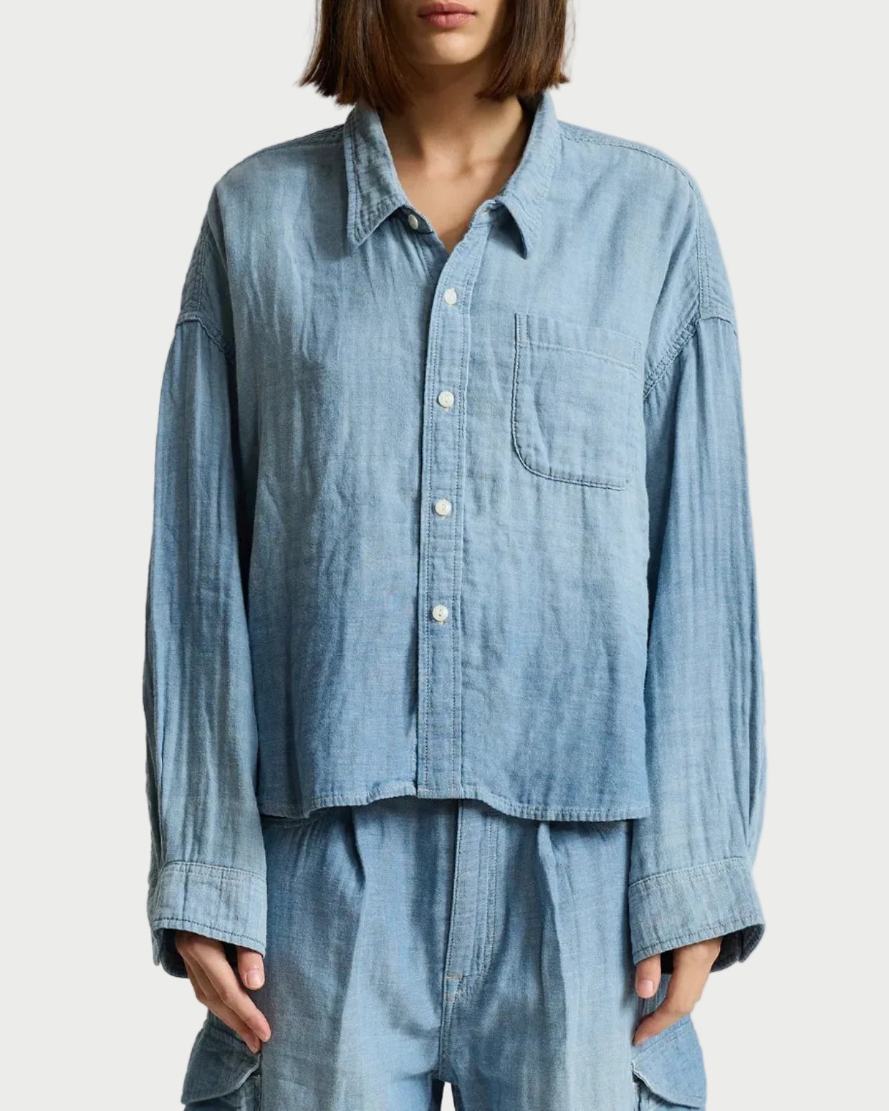 A model wearing a light blue chambray button-up shirt and matching light blue trousers, photographed against a neutral light gray studio background.